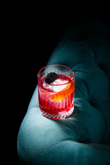 red cocktail in a transparent glass with round ice, whipped cream and black berry on top, side view