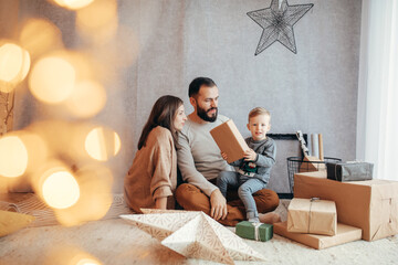 In a beautifully decorated home glowing with festive lights, a young family gathers around,...