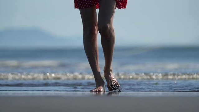 Female bare feet walking on wet black sand on sandy beach of Pacific Ocean. Low section woman body part, long and slender stepping woman legs. Model in red polka dot short dress. Slow motion, handheld