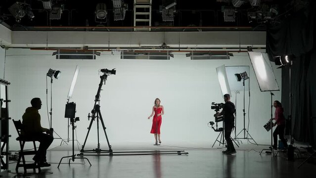 Professional filming pavilion with a white cyclorama. Shooting of a girl in a red dress who sings into a retro microphone. Director, Cameraman and crew in Backstage.