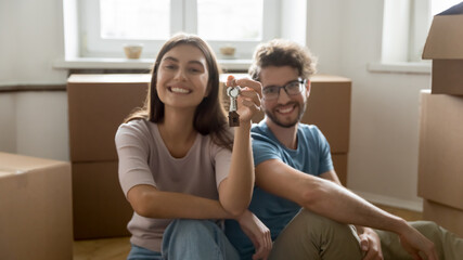 Young couple sit on floor near heap of carton boxes with personal stuff smile look at camera showing keys from their new bought or rented flat, start independent life together. Tenancy, cohabitation