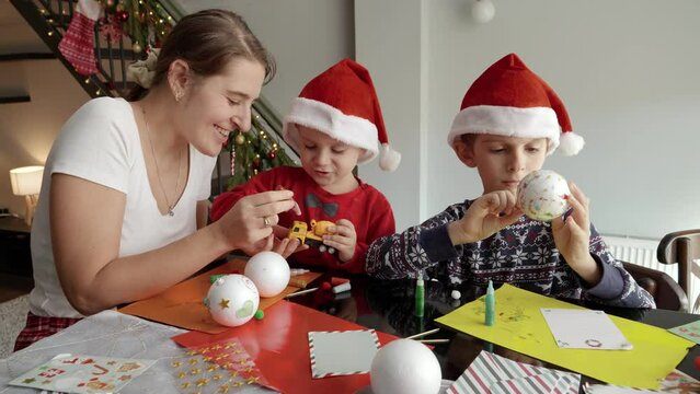Kids with mother painting and decorating Christmas baubles in living room. Winter holidays, family time together, kids with parents celebrating