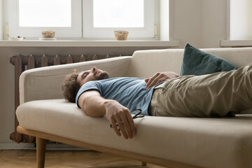 Tired young man having daytime nap on sofa at home. Overloaded guy in casual wear falling asleep on...