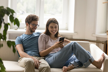 Happy young married couple spend time at home sitting on couch use smartphone together, looking at screen watch video, shopping online. Loving man hug smiling woman enjoy spend time in social networks