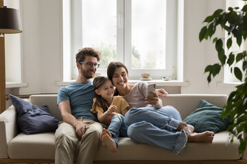 Smiling young spouses sit on sofa with little daughter hold smartphone, take self-portrait on...