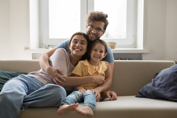 Loving young man hug wife and little daughter sitting on sofa, happy family smile pose looking at camera. Happy parents spend weekend time with cute kid at home, enjoy time together. Parenthood, love