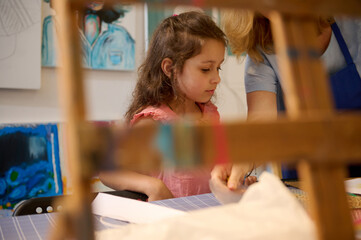 View though a blurred wooden easel of a cute little kid girl learning painting and art in a...