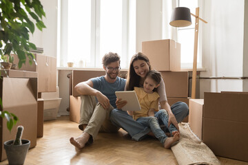 Young family with child sit on floor at new flat near boxes with stuff use tablet, browse internet, discuss furniture purchase. Spouses buy items for new house on relocation. Move-in day, e-commerce - obrazy, fototapety, plakaty