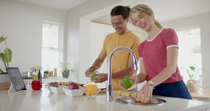 Happy diverse couple preparing and washing fresh vegetables in kitchen with tablet, slow motion