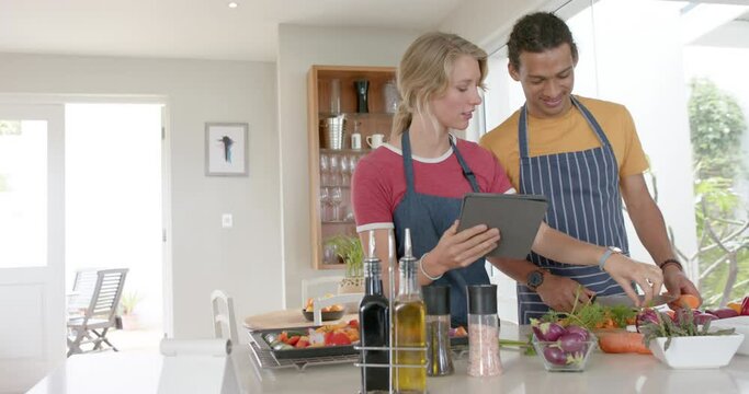 Happy diverse couple preparing fresh vegetables and using tablet in kitchen, slow motion