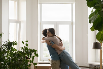 Overjoyed loving young spouses cuddling standing in living room with cardboard boxes, congratulate...