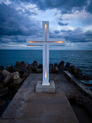A large Christian cross stands at the edge of a pier against a dramatic sky and sea, seen from above