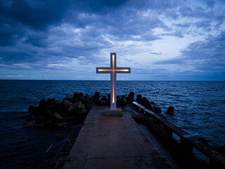 A large Christian cross stands at the edge of a pier against a dramatic sky and sea, seen from above - obrazy, fototapety, plakaty