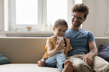 Happy young man sit on cozy couch with little daughter use mobile phone, communicate, play online...
