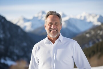 Portrait of a grinning man in his 50s wearing a classic white shirt against a snowy mountain range....