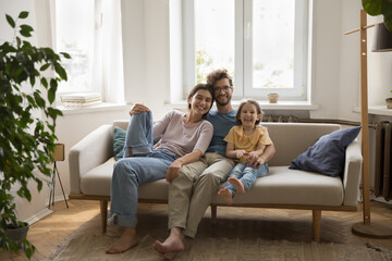 Young happy family sit on sofa in living room looking at camera, pose, spend weekend time together. Caring spouses enjoy friendly relationship, express love and tenderness. Unity, parenthood, devotion
