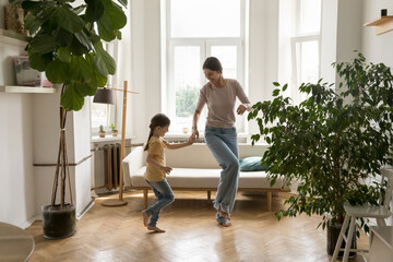 Beautiful mother teach cute little kid to dance, dancing to favorite music in living room, enjoy active weekend at home. Daughter and mom holding hands, moving in cozy flat. Motherhood, hobby, fun