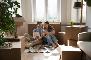 On relocation day happy young wife and husband sit on floor giving high five, boxes with belongings and heap of color samples tiles, renovation brochures nearby. Repair works, renovation, new house - obrazy, fototapety, plakaty