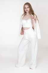 Woman in white suit jacket, trousers and slingbacks standing on white background. Blondie fashion model holding straps of corset top with fingers of one hand and belt loop on trousers with other hand