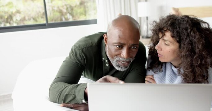 Happy mature diverse couple lying on bed using laptop at home, slow motion