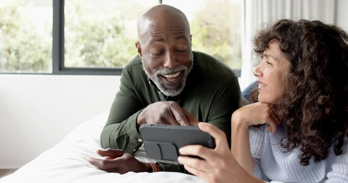 Happy mature diverse couple lying on bed using smartphone at home, slow motion