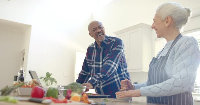 Happy diverse senior couple preparing vegetables and using tablet in kitchen, slow motion