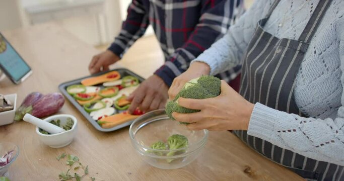 Happy diverse senior couple preparing vegetables and using tablet in kitchen, slow motion