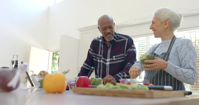 Happy diverse senior couple preparing vegetables, using tablet in kitchen, copy space, slow motion