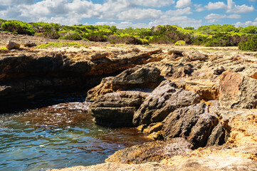 Stone coast with a small bay in front oft the green forest under a blue cloudy sky.