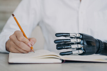 Focused individual writing in notebook with a mechanical prosthetic hand, showcasing adaptive...