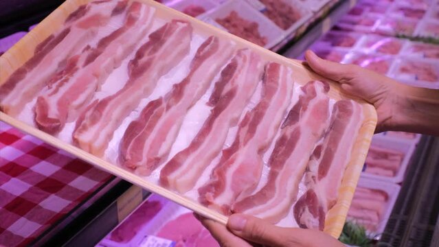Shopper in store carefully examines package of sliced bacon. Coming to store, person is faced with variety of options when choosing bacon. Buyer is betting on quality and taste of fresh bacon.