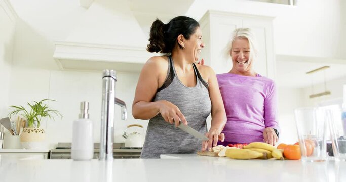 Two happy diverse senior women chopping fruits and laughing in sunny kitchen, slow motion