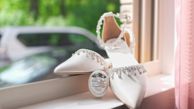 A beautiful shot of wedding shoes laying on the window sill with rings at the base of the shoes. Pink curtains are near the shoes