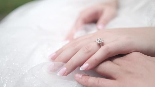 A 4k close up shot of a brides hand wearing a sparkly wedding ring. The bride is wearing a white wedding dress and  her nails have a french manicure. Her hand is resting on top of the grooms.