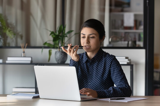 Serious confident Indian business woman, leader working at laptop, recording audio message on smartphone, using voice recognition app on mobile phone in office, giving virtual audio command