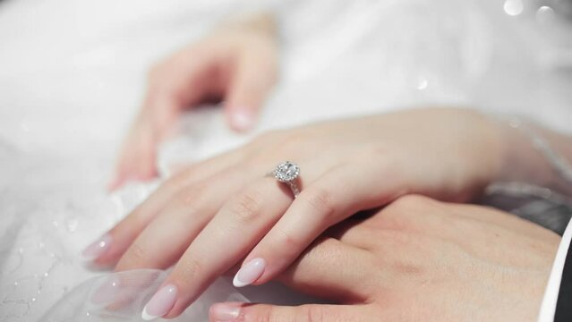 A close up shot of a brides hand wearing a sparkly ring resting on top of the grooms hand. The bride is wearing a white wedding dress. Brides nails are done in a french manicure.