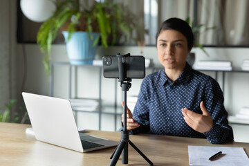 Serious Indian presenter girl, speaker talking at webcam on smartphone. Online teacher, tutor, coach giving webinar from home. Woman using mobile phone on tripod for remote online conference call - obrazy, fototapety, plakaty