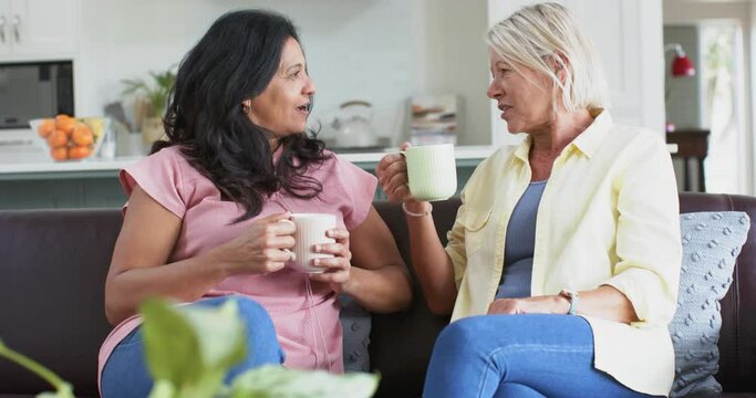 Happy diverse senior women with coffee, discussing on sofa in sunny living room, slow motion