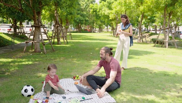 Boy practices learning to draw and paint next to him. father is sitting on grass using  laptop computer to work online. Caucasian family of father, mother and son sitting and relaxing in village park.