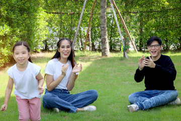 Fototapeta na wymiar Happy family having fun together in summer garden, cute daughter girl running around parents who sitting on green grass, kid playing with her father and mother at backyard. Family love bonding.
