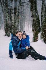 Fototapeta na wymiar Laughing dad with a little squinting girl on his knees sits under a snowfall in the forest