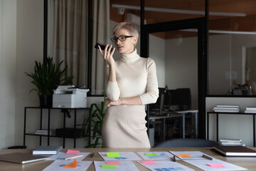 Successful stylish businesswoman talking on mobile phone on speaker, giving voice command to virtual assistant, recording message at workplace with scrum arranged paper stickers and documents