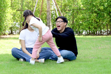 Fototapeta premium Happy family having fun together in summer garden, cute daughter girl running around parents who sitting on green grass, kid playing with her father and mother at backyard. Family love bonding.