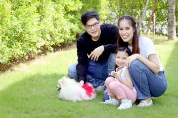 Obraz premium Happy family with pet has fun together in summer green garden. Father, mother and kid play with fluffy white Pomeranian dog, mom hugging cute daughter girl, joyful parents with child at backyard.