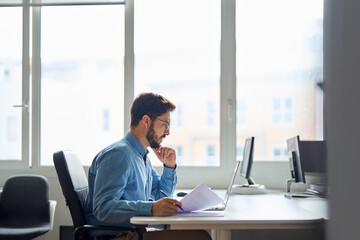 Busy serious young business man working on laptop computer holding documents at workplace. Latin...