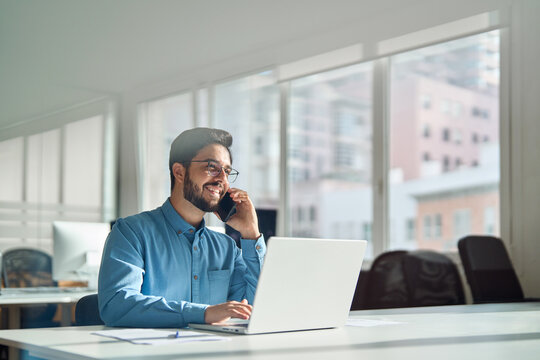 Young happy busy latin businessman talking on phone using laptop computer in office. Smiling hispanic business man making call on mobile cellphone, consulting client or having work cell conversation.