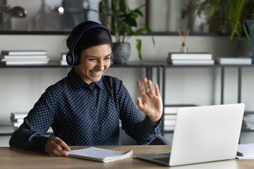 Happy Indian student girl wearing headphones, making video call to teacher, talking to tutor, waving hello at webcam, looking at screen, smiling, writing notes. Woman attending virtual conference chat - obrazy, fototapety, plakaty