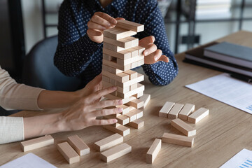 Business team colleagues playing jenga board game together, holding tower building, high stack of wooden blocks from falling, dealing with challenge, risk, teamwork and teambuilding skills. Close up - obrazy, fototapety, plakaty