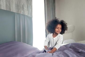 Cute little African girl child with black curly hair making a bed after waking up in morning at...