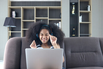 Happy smiling beautiful teenage girl with curly hair wearing headphones, using laptop computer for practicing to sing song while sitting on sofa in home living room.